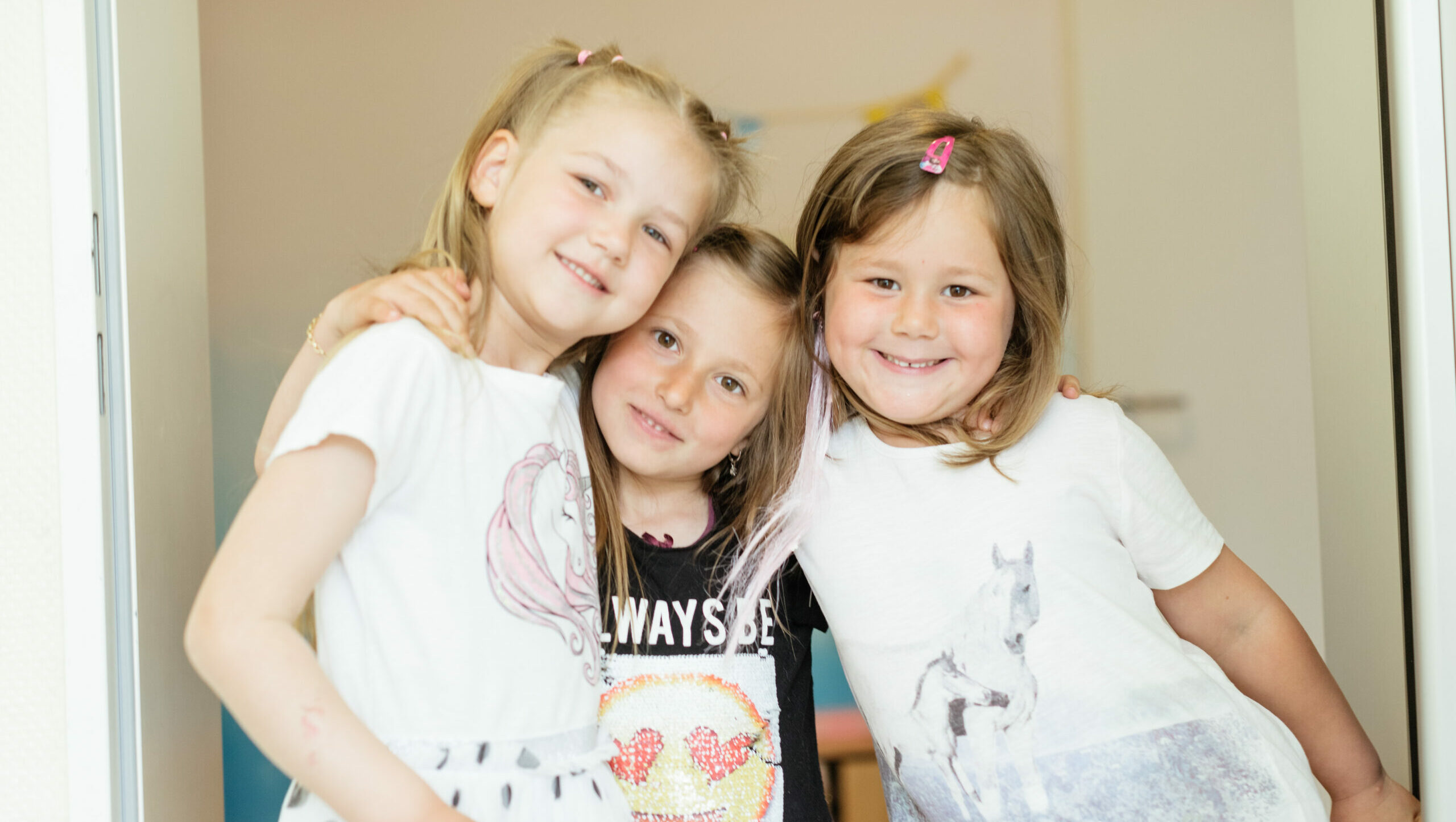 Drei kleine Mädchen stehen lächelnd in die Kamera und nehmen sich in die Arme.