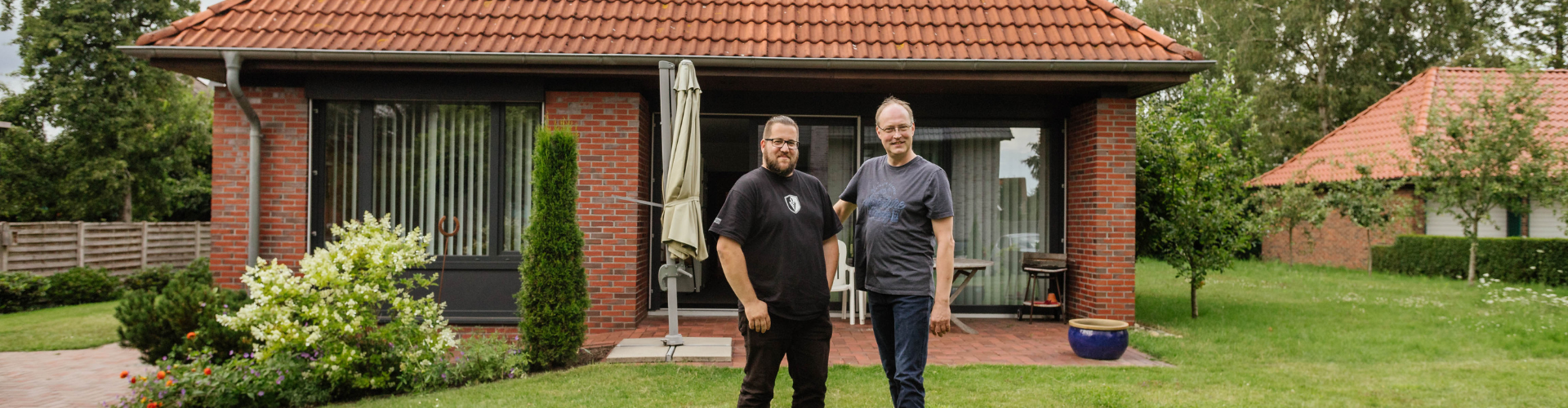 Zwei Männer stehen nebeneinander vor einem Haus auf einer grünen Rasenfläche und lachen in die Kamera.