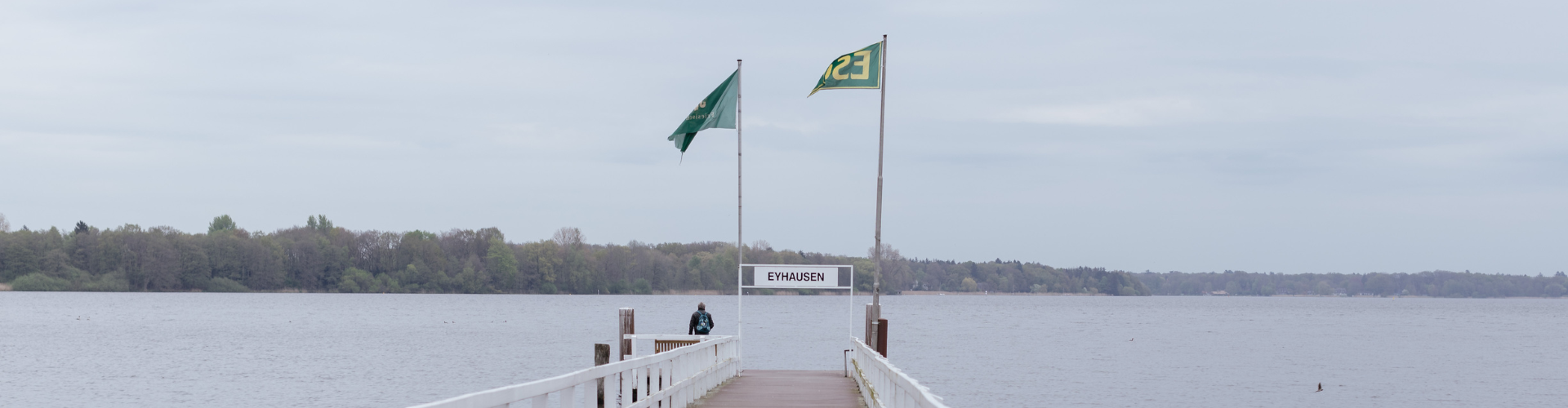 Draußen am Meer. Man sieht im Hintergrund einen Wald und einen langen Steg am Wasser. Auch sieht man zwei Flaggen und ein Schild mit der Aufschrift "Eyhausen"