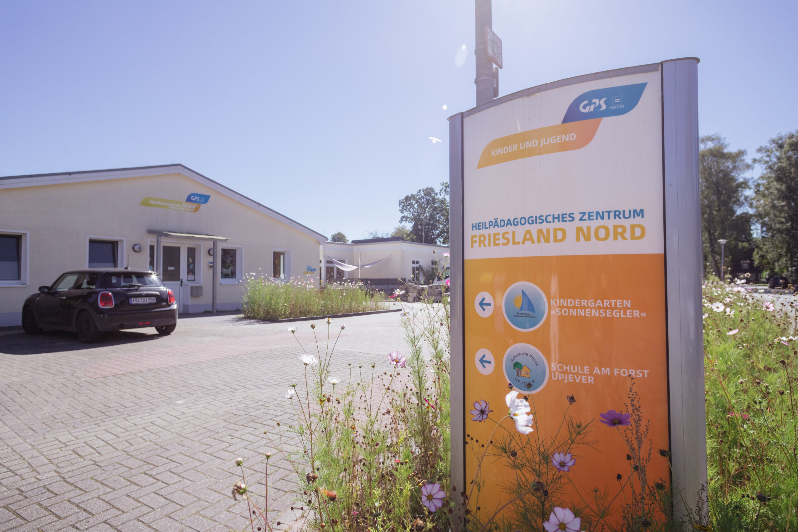 Draußen bei sonnigen Wetter sieht man die Einrichtung des Heilpädogischen Zentrums Friesland Nord. An der Straße ist eine Infotafel zum "Kindergarten Sonnensegler" und die "Schule am Forst Upjever".
