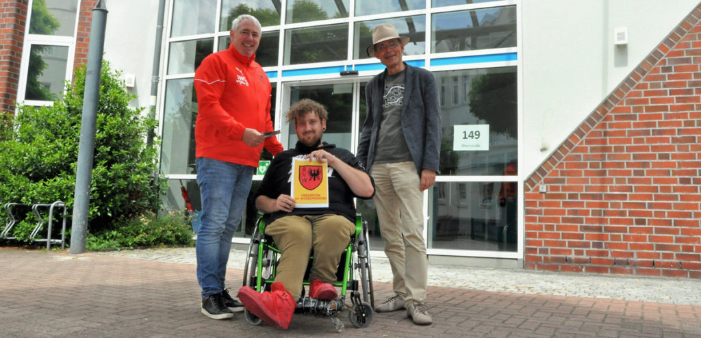 Man sieht 2 ältere Männer und 1 junger Mann im Rollstuhl, der ein Blatt zeigt mit dem Wappen des Fußballvereins SV Wilhelmshaven