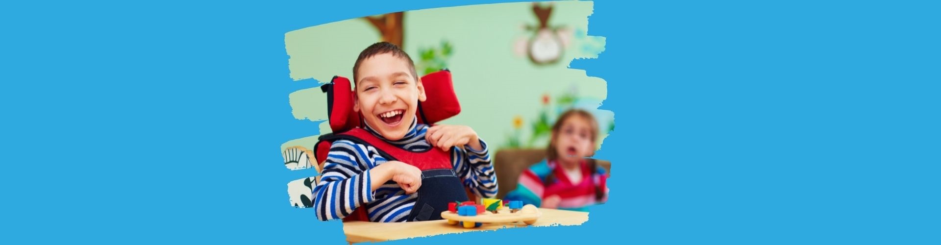 Eine Junge mit einer Behinderung im Rollstuhl. Er sitzt an einem Tisch mit Spielsachen. Im Hintergrund sieht man ein junges Mädchen mit eine Behinderung