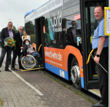 Man sieht Mitarbeiter der GPS, den Geschäftsführer Klaus Puschmann mit einem Blumenstrauß, eine junge Frau im Rollstuhl vor einem Bus an der Straße. Dieser Bus steht an der neuen Bushaltestelle der GPS. Der Busfahrer steht vor dem Vordereingang des Busses und schaut auch lächelnd in der Kamera. Der Geschäftsführer der Stadtwerke steht mit der jungen Frau im Rollstuhl vor dem letzten Hintereingang mit einer Rampe.