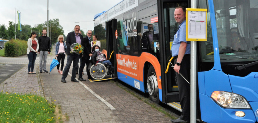 Man sieht Mitarbeiter der GPS, den Geschäftsführer Klaus Puschmann mit einem Blumenstrauß, eine junge Frau im Rollstuhl vor einem Bus an der Straße. Dieser Bus steht an der neuen Bushaltestelle der GPS. Der Busfahrer steht vor dem Vordereingang des Busses und schaut auch lächelnd in der Kamera. Der Geschäftsführer der Stadtwerke steht mit der jungen Frau im Rollstuhl vor dem letzten Hintereingang mit einer Rampe.