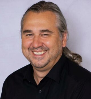 Markus Haarmann. Eine lächelnder Mann mit langen grauen Haaren mit einem Zopf gebunden. Er trägt einen Bart.