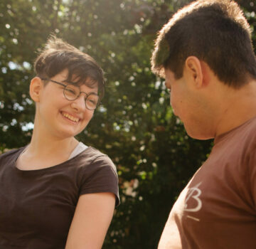 Eine junge Frau und ein junger Mann sind draußen im Garten. Sie lächeln sich an.