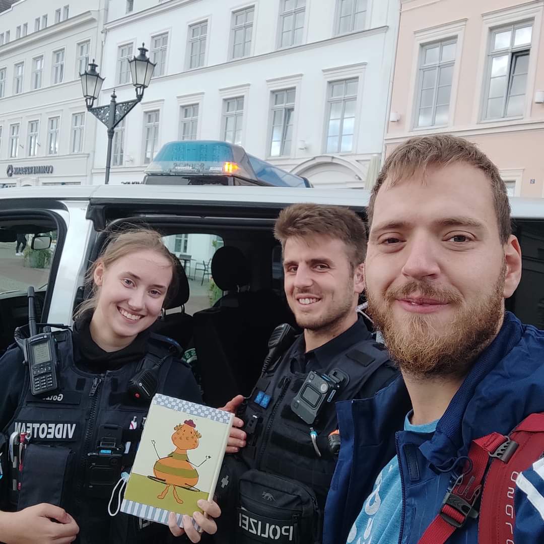 Ein junger Mann namens Jan Kleen, ein Polizeibeamter und eine Polizeibeamtin stehen vor ein Polizeiauto. Sie schauen freundlich in die Kamera. Eine Polizistin hält ein Kinderbuch.