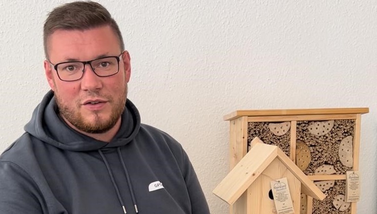 Ein GPS Mitarbeiter namens Marcel Menze steht vor der Kamera. Er schaut freundlich. Im Hintergrund ist ein Vogelhaus und ein Insektenhotel aus Holz zu sehen.