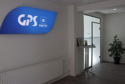 Ein Flur in einer GPS Einrichtung auf dem Bild. An der Wand ist oben ein blau leuchtendes Schild "das GPS Logo". Auf einem Tisch sind auch verschiedene Flyer.