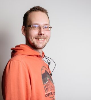 Dennis Kauffmann Bürohelfer Unternehmenskommunikation Mann im orangenen Pullover lacht