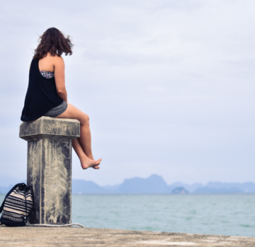Eine junge Frau sitzt draußen auf einem Anleger aus Stein und schaut aufs Meer.