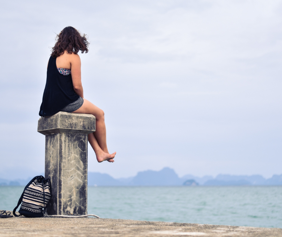 Eine junge Frau sitzt draußen auf einem Anleger aus Stein und schaut aufs Meer.
