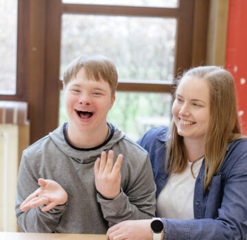Ein Junge mit einer Behinderung und eine junge Frau sitzen an einem Tisch. Sie lächeln.