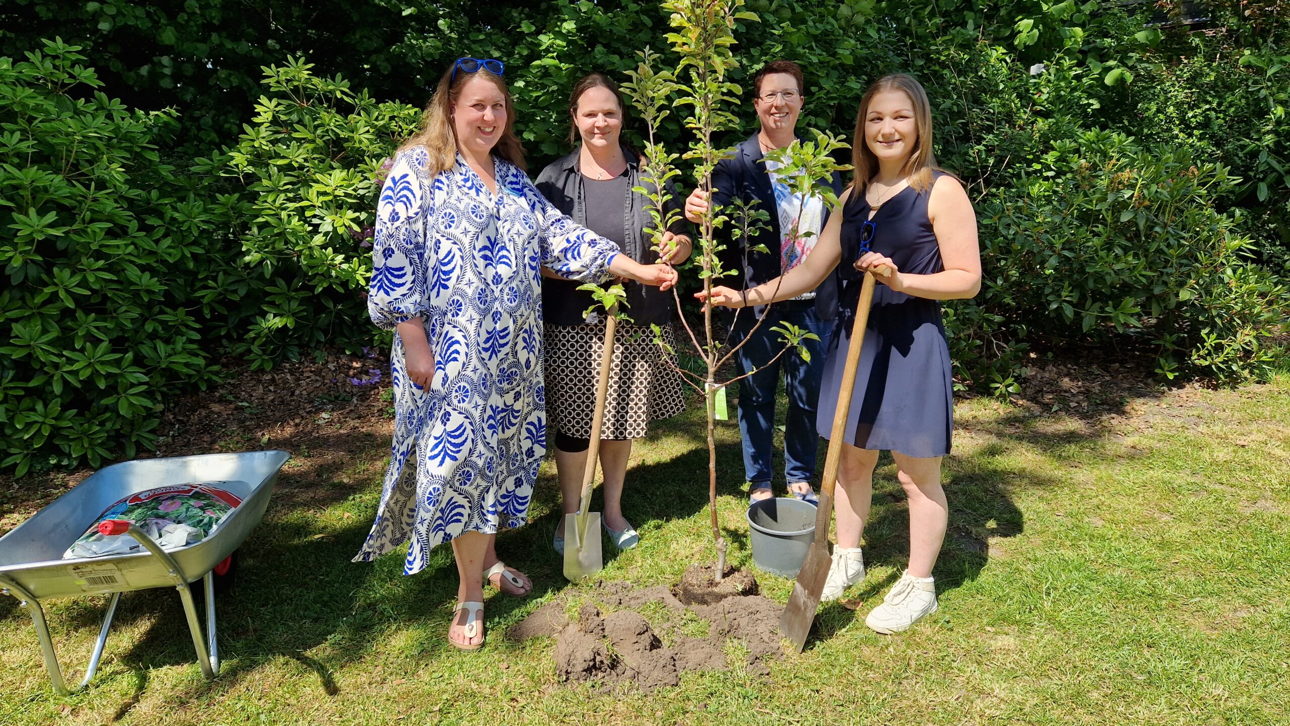 Vier GPS Mitarbeiterinnen stehen draußen im Garten und pflanzen gerade einen kleinen Baum ein.