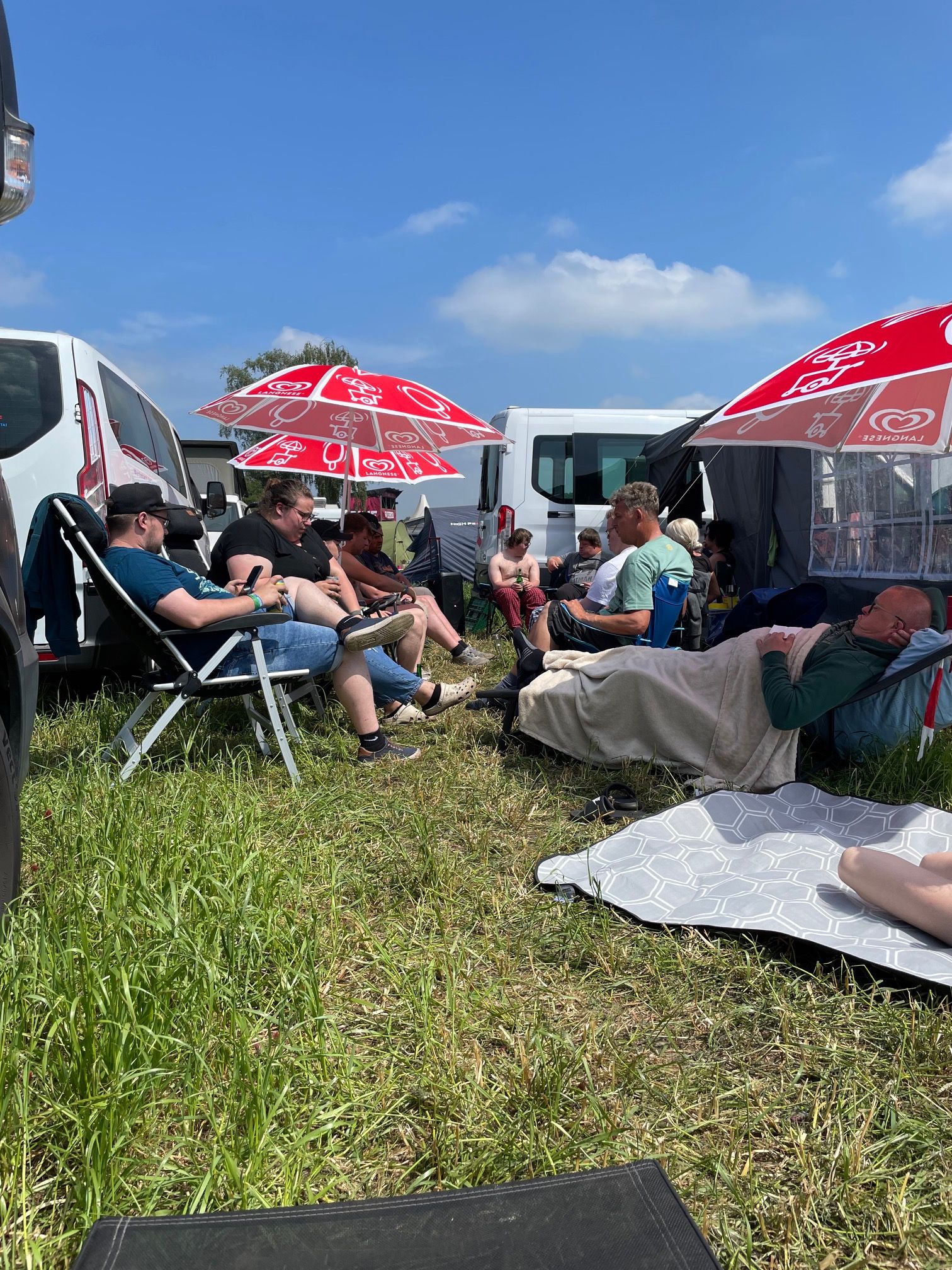 Eine Gruppe Menschen sitzen auf Stühlen und Liegen. Man sieht auch Autos und Sonnenschirme.