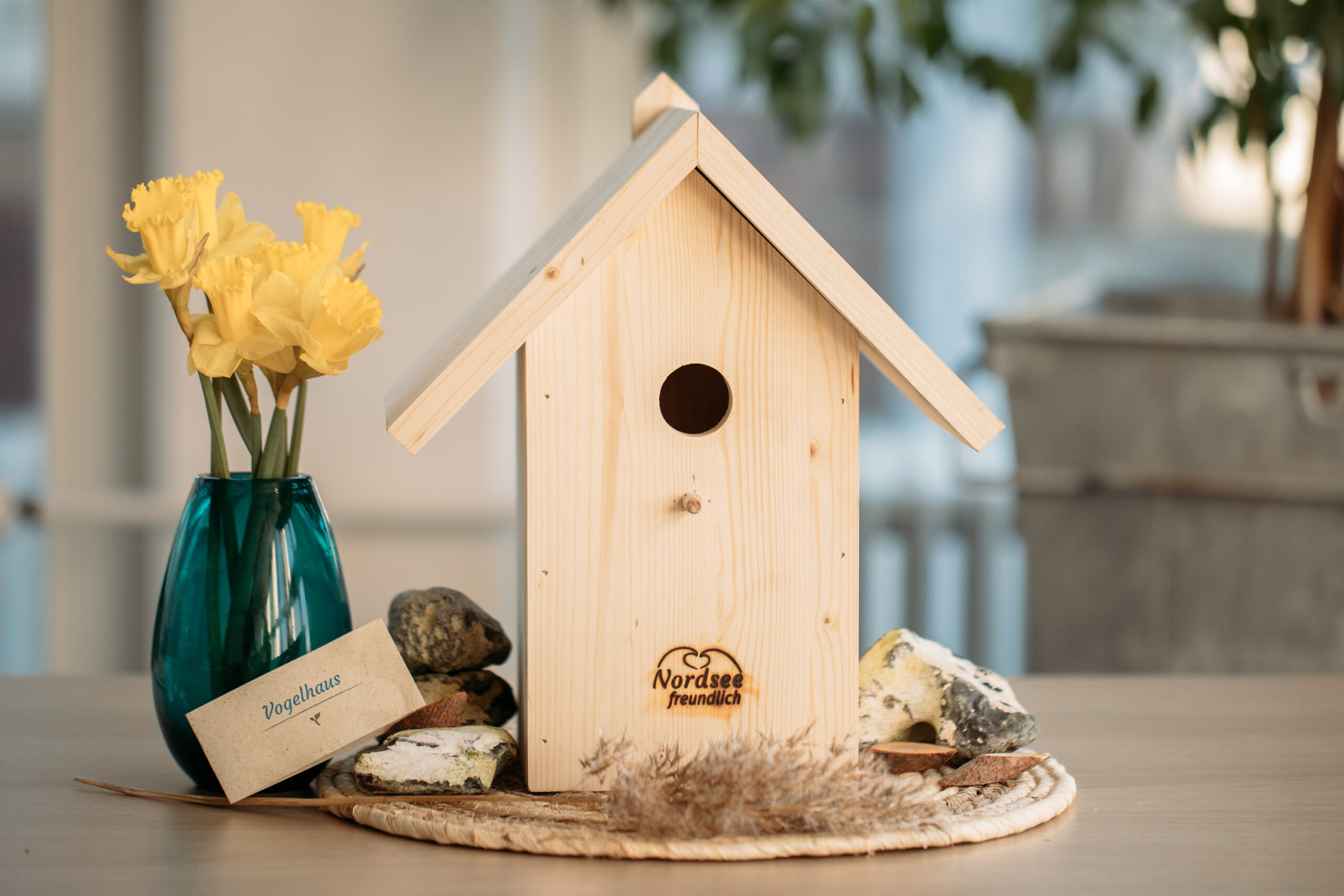 Ein selbst gebautes Vogelhäuschen mit kleinen Steinen, eine Pflanze und eine gelbe Blume in einer blauen Vase. Auf dem Vogelhäuschen ist das Logo "Nordseefreundlich" und eine Karte "Vogelhaus" abgebildet.