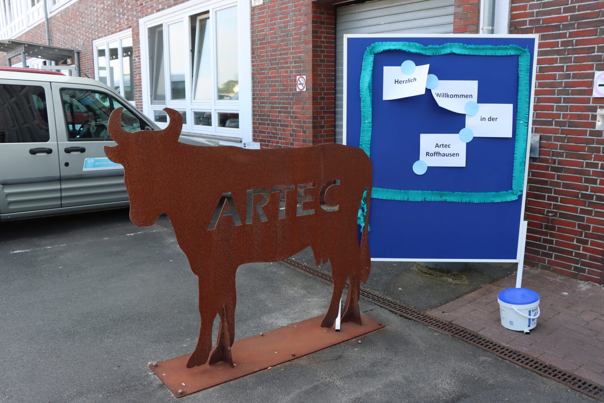 Draußen vor dem Eingang der GPS ARTEC Roffhausen steht ein selbst gebauteter Stier aus Metall. Neben dem Stier ist eine Infotafel, wo daran auf Zetteln steht "Herzlich Willkommen in der Artec Roffhausen".