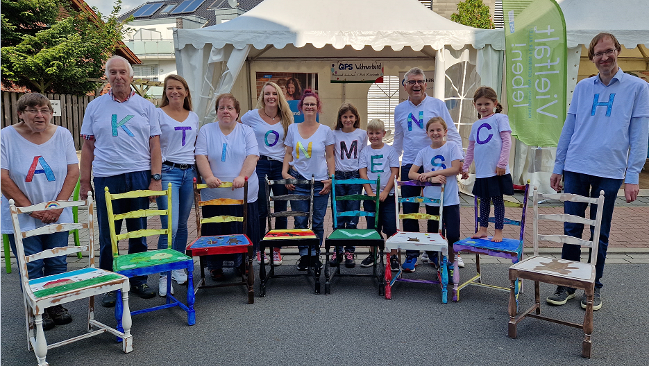 Kinder und Erwachsene stehen draußen vor farbigen Stühlen. Auf ihren T-Shirts steht ein Buchstabe. Zusammen sieht man und liest man das Wort "Aktion Mensch"
