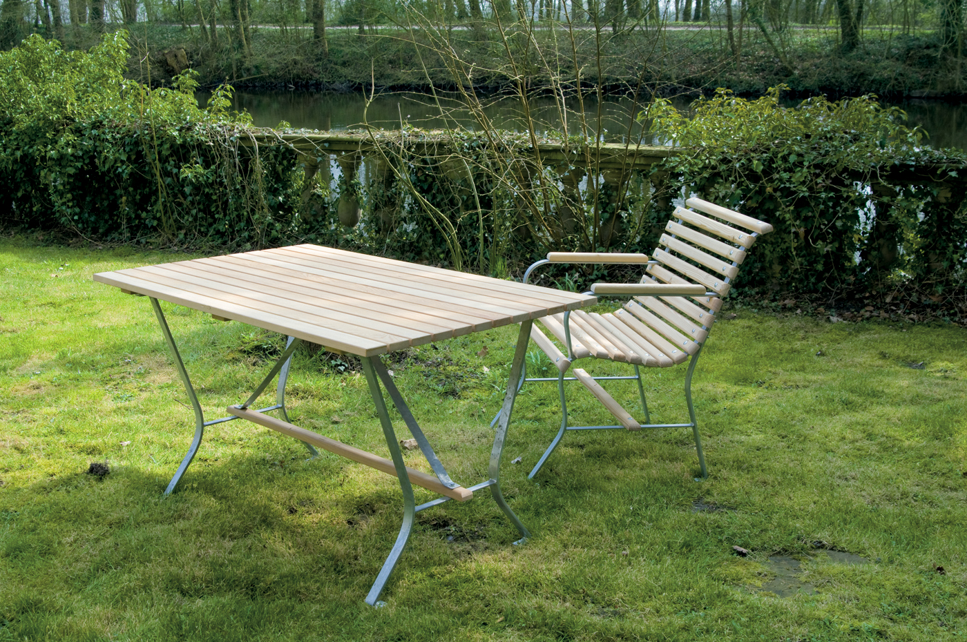 Draußen im einem Garten sieht man einen Tisch und einen Stuhl aus Holz. Im Hintergrund sieht man einen Fluss und eine große lange Hecke.
