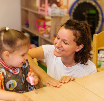 Eine junge Erzieherin und ein kleines Mädchen lächeln sich gegenseitig an. Sie sitzen an einem Tisch.