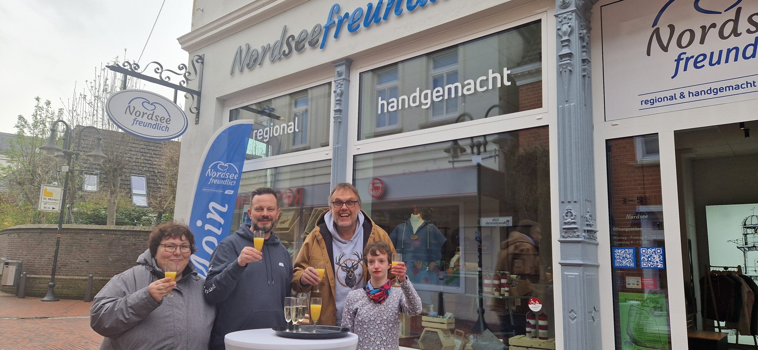 Geschäftsführer von der GPS Klaus Puschmann, ein GPS Mitarbeiter und zwei Frauen sind draußen vor dem GPS Laden "Nordseefreundlich". Alle stoßen mit einem Glas Orangensaft auf ein bestehendes Jahr des GPS Ladens "Nordseefreundlich".