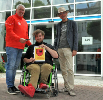 Man sieht 2 ältere Männer und 1 junger Mann im Rollstuhl, der ein Blatt zeigt mit dem Wappen des Fußballvereins SV Wilhelmshaven