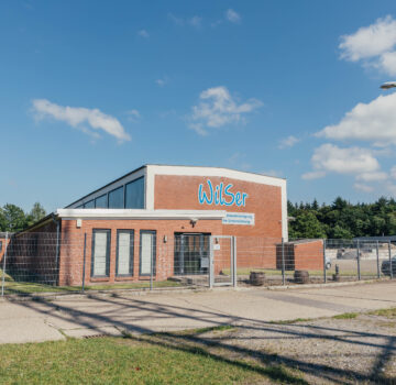 Ein Gebäude mit einer großen Halle. Die GPS Einrichtung "WilSer". Die Sonne scheint draußen. Parkplatz mit Autos.