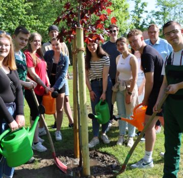 Eine Gruppe von FSJlern stehen vor dem gepflanzten Baum. Sechs von ihnen haben eine Schaufel und eine Gießkanne