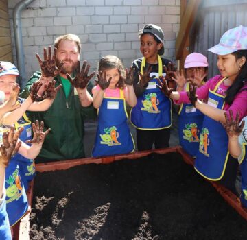 Ein Mann (Gärtner) mit acht kleinen Kindern stehen vor einem Beet mit Sand. Sie halten alle ihre schmutzigen Hände hoch.