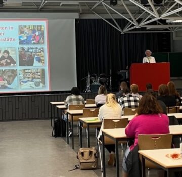 Ein großer Saal mit vielen Leuten. Es wird gerade eine Rede gehalten. Auf einer Leinwand werden Bilder zum Thema "Arbeiten in der Förderstätte" gezeigt.