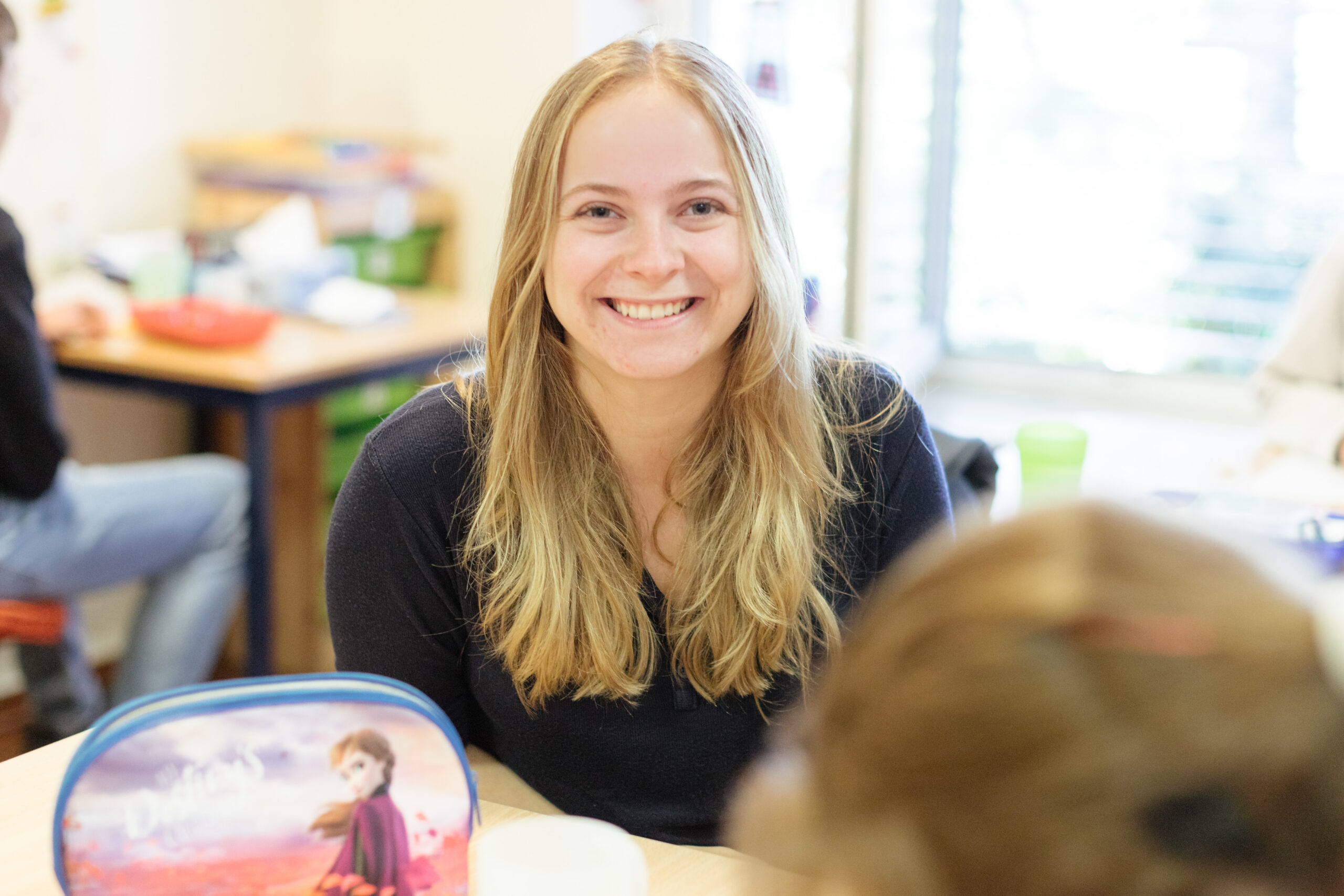 Eine junge Frau sitzt an einem Tisch. Sie ist mit anderen Kindern in einem Klassenraum. Sie lächelt.