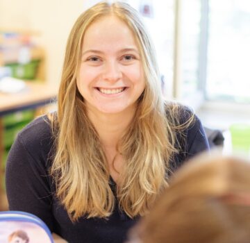 Eine junge Frau sitzt an einem Tisch. Sie ist mit anderen Kindern in einem Klassenraum.