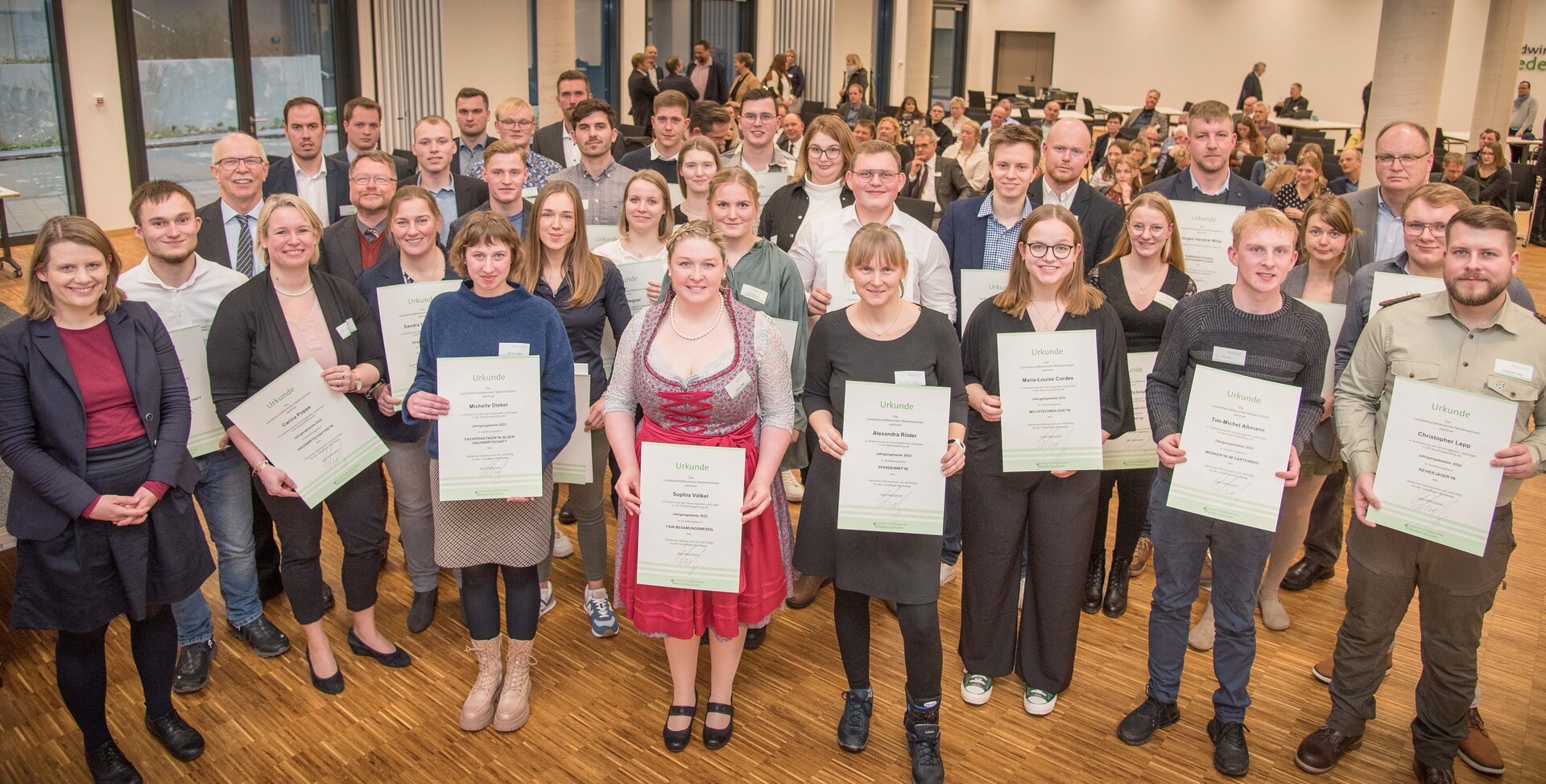 Viele junge Leute bei einer Auszeichnungsfeier zu ihrer Werker-Ausbildung. Sie halten alle Ihre Urkunden in der Hand. Hinten sieht man auch das Publikum.