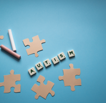 Kleine braune Puzzleteile aus Papier, zwei verschiedene, farbige Wachsmalstifte und jeweils sechs weiße kleine Würfel mit Buchstaben. Die weißen Würfel ergeben das englische Wort für "Autism", das auf Deutsch "Autismus" heißt.