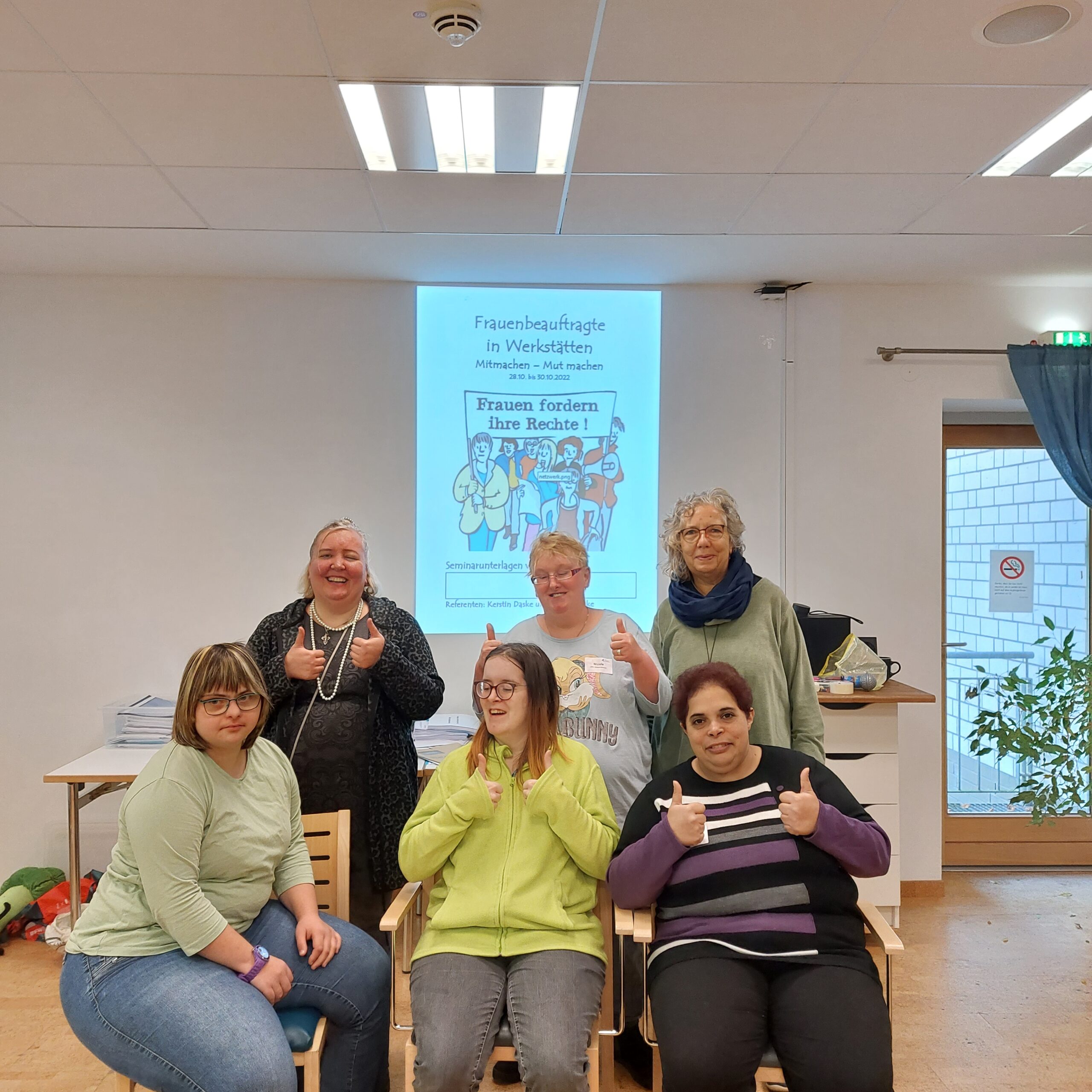 Frauenbeauftragte Andrea Hicken und fünf GPS Beschäftigte sind in einem Raum. Sie stehen oder sitzen. Im Hintergrund an der Leinwand sie man ein Bild mit dem Thema "Frauenbeauftragte in Werkstätten - Frauen fördern ihre Rechte!" Sie halten alle ihre Daumen hoch.