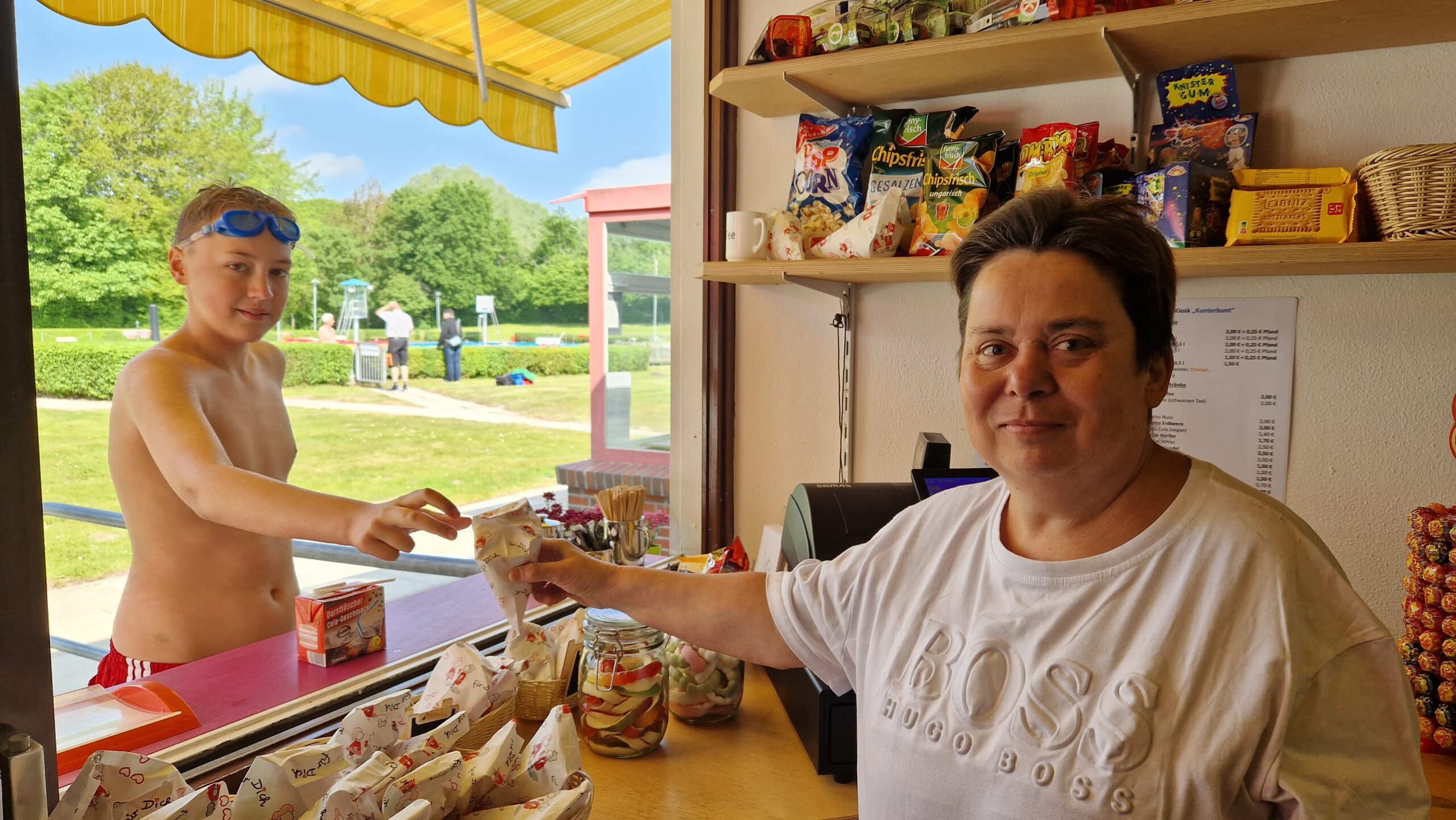 Man sieht drinnen den Kiosk Kunterbunt. Eine junge Frau gibt einen Jungen eine Tüte mit Süßigkeiten.