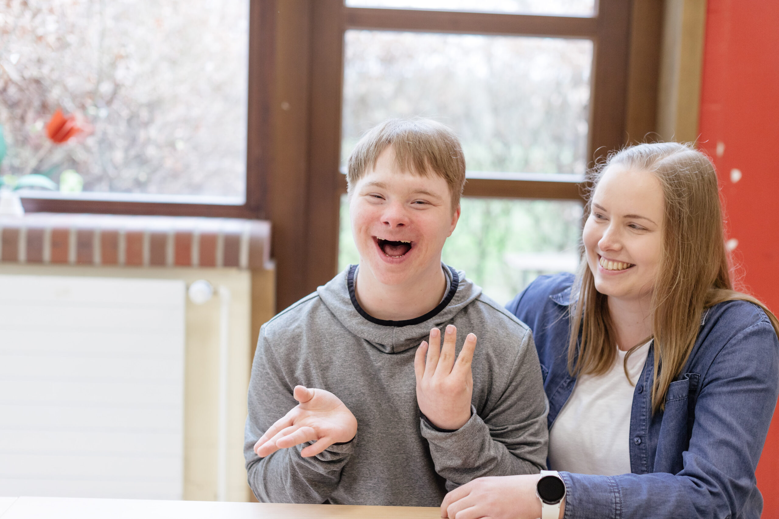 Ein Junge mit einer Behinderung und eine junge Frau sitzen an einem Tisch. Sie lächeln.