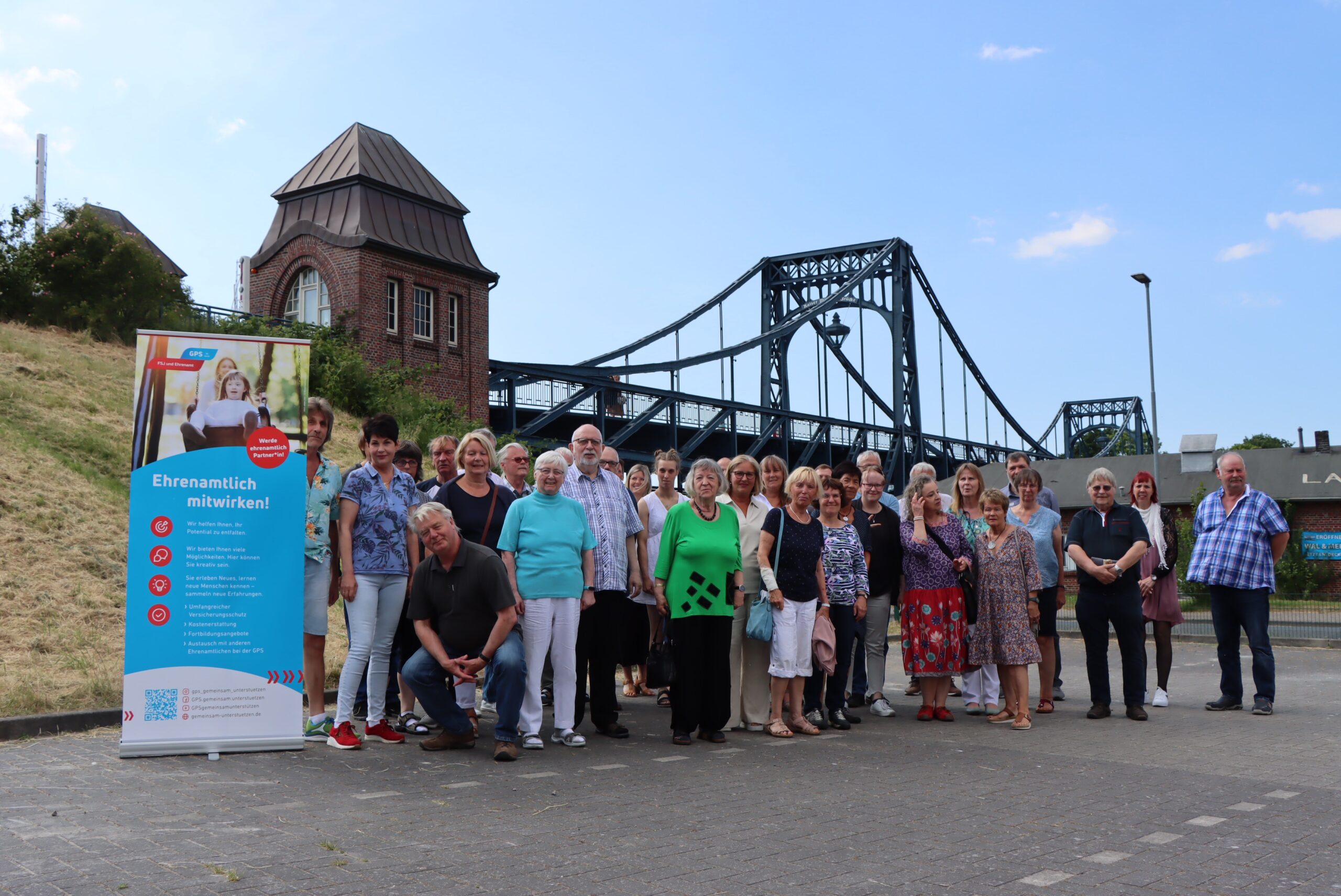 Eine Gruppe von Ehrenamtlichen und GPS Mitarbeiter sind draußen auf einem Parkplatz beim Südstrand. Im Hintergrund sieht man die Kaiser-Wilhelm-Brücke.