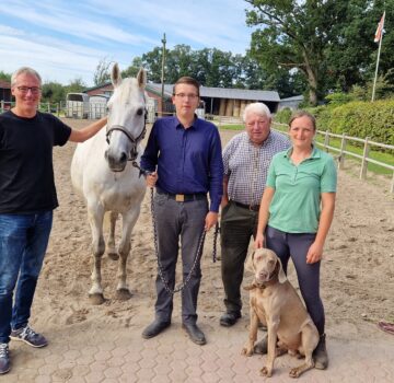 Drei Männer und eine Frau mit einem Pferd und einem Hund draußen auf einem sandigen Platz. Sie schauen alle in die Kamera.