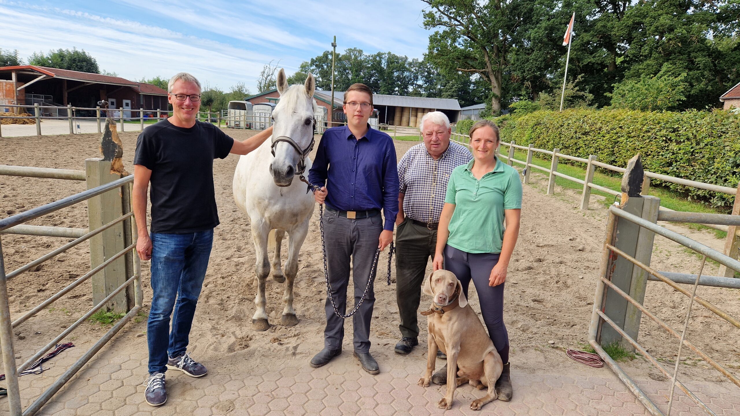 Drei Männer und eine Frau mit einem Pferd und einem Hund draußen auf einem sandigen Platz. Sie schauen alle in die Kamera.