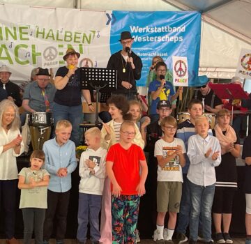 Kinder und Erwachsene in einem Zelt. Man sieht ein paar Beschäftigte der GPS auf der Bühne. Sie machen Musik. Im Hintergrund sieht man ein großes Plakat auf dem darauf steht "Keine Halben Sachen von der Werkstatt Westerscheps".