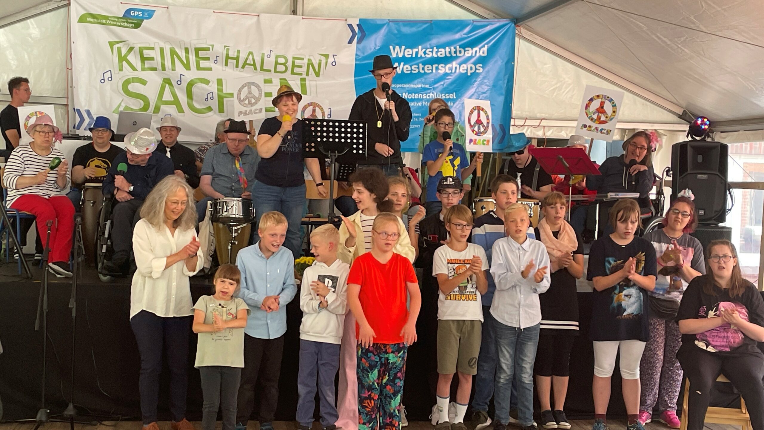 Kinder und Erwachsene in einem Zelt. Man sieht ein paar Beschäftigte der GPS auf der Bühne. Sie machen Musik. Im Hintergrund sieht man ein großes Plakat auf dem darauf steht "Keine Halben Sachen von der Werkstatt Westerscheps".