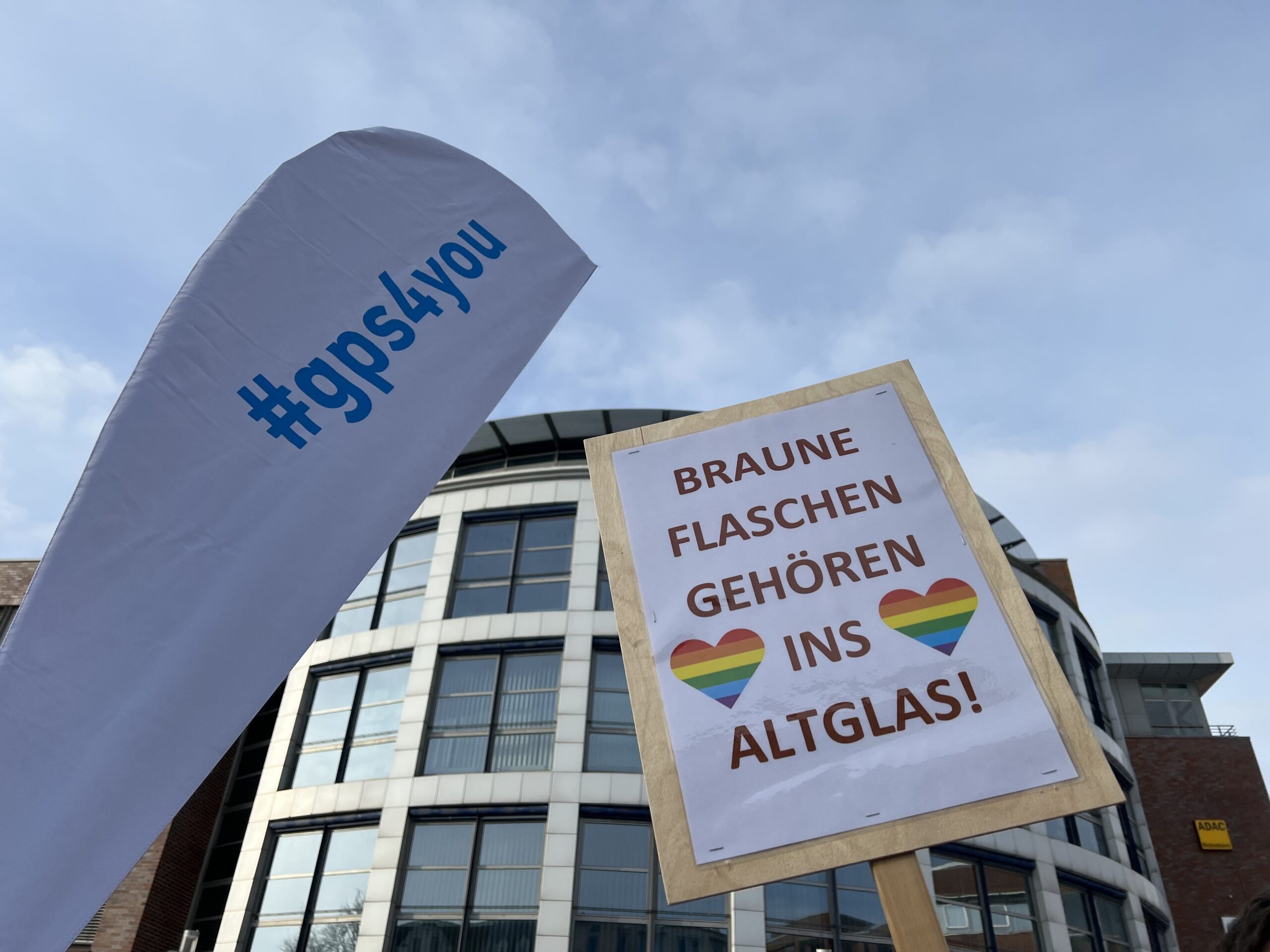 Draußen bei einer Demonstration gegen Ausgrenzung und Rechtsradikalismus vor einem Gebäude. Man sieht eine "gps4you" Flagge und ein Plakatschild mit der Aufschrift "Braune Flaschen gehören ins Altglas"