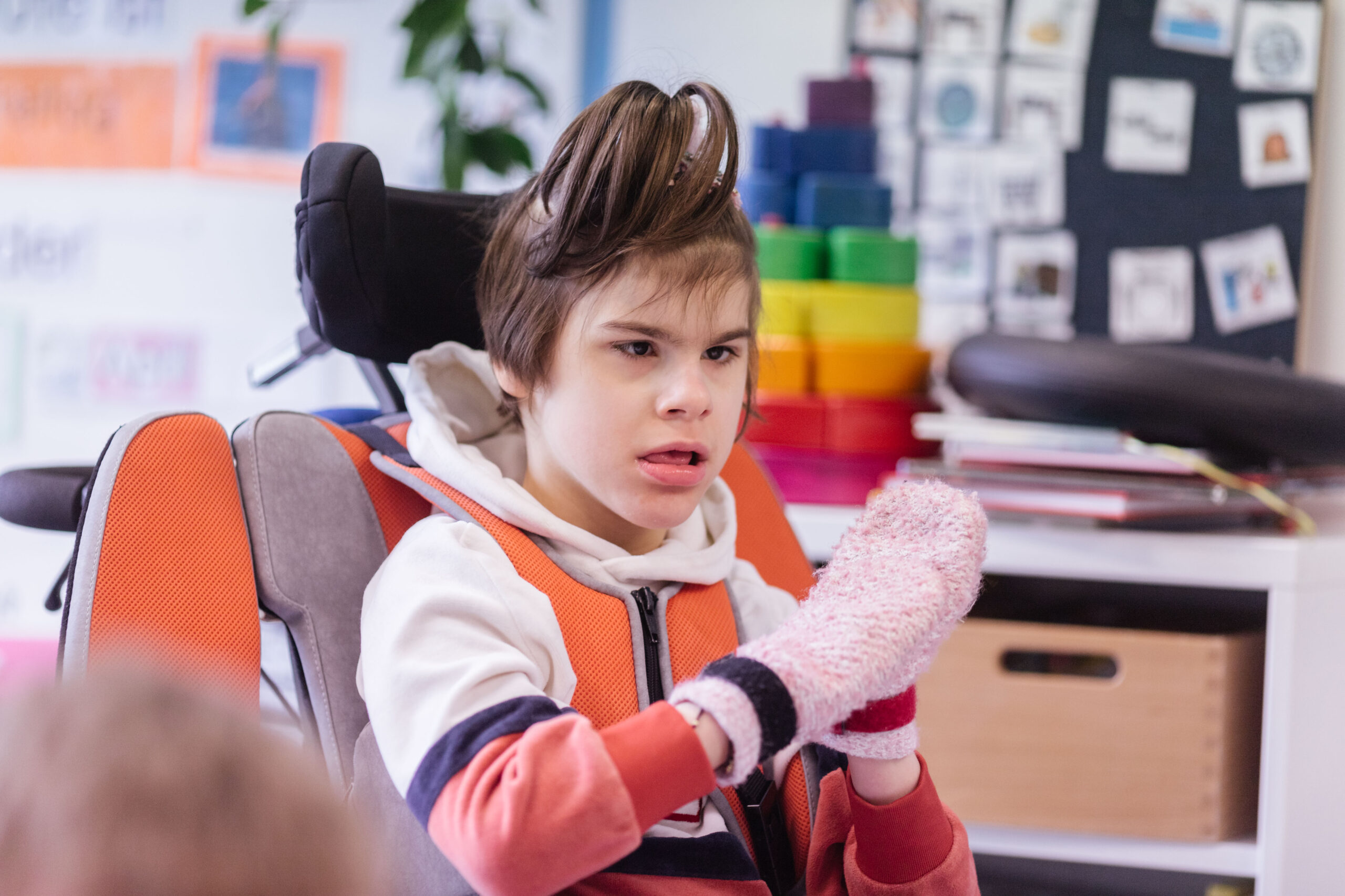 Eine Schülerin mit einer Behinderung sitzt in einem Rollstuhl. Sie hat farbige Handschuhe an und hält ihren Hände zusammen.