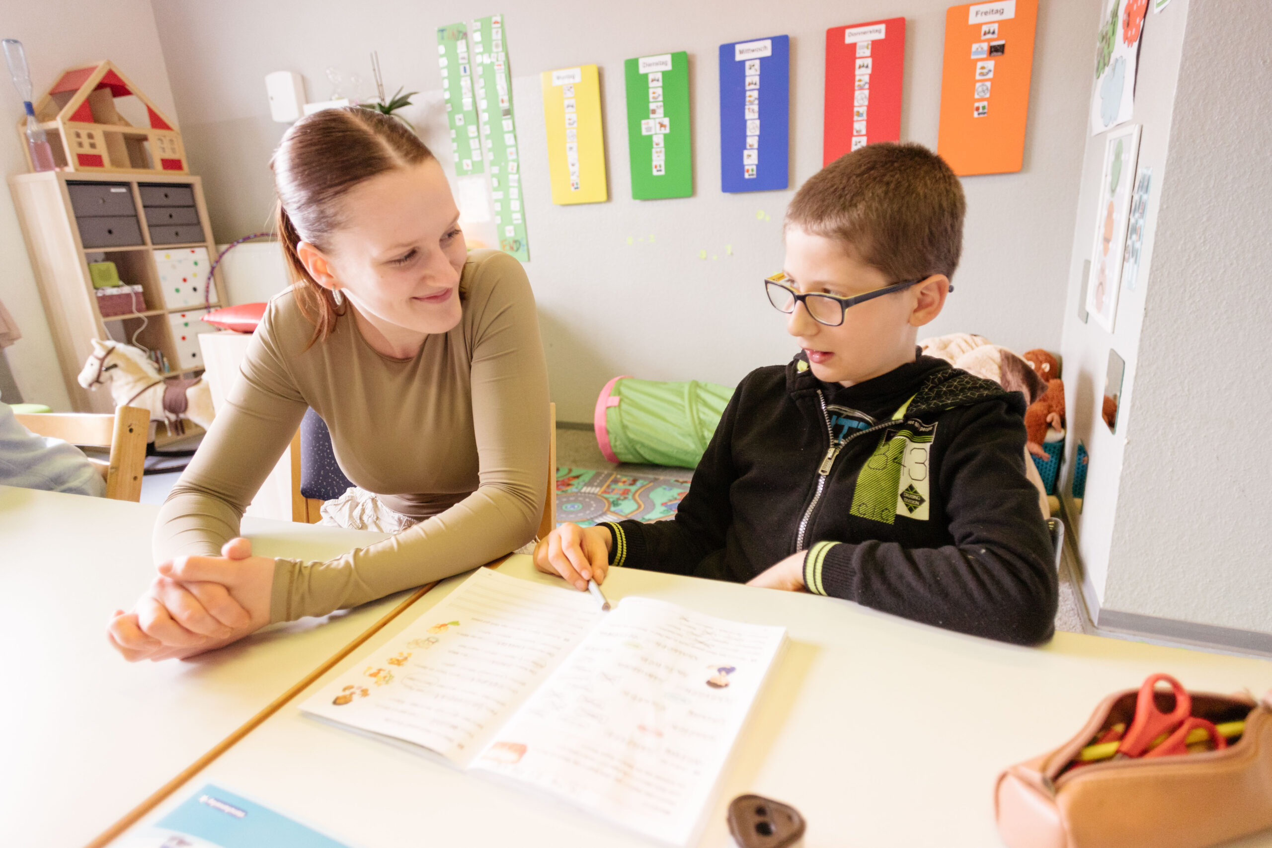 Eine junge Lehrerin hilft einem Schüler bei einer Aufgabe. Sie schauen sich gegenseitig lächelnd an.