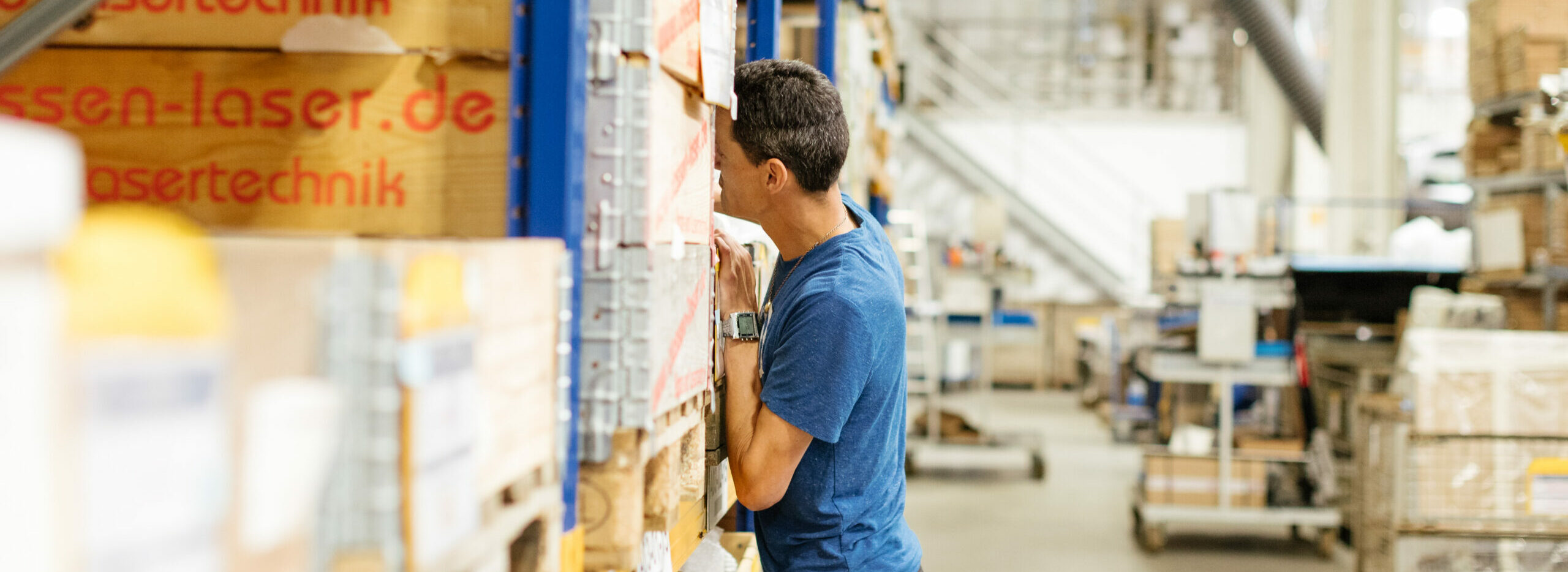 Ein Mann ist in einer großen Halle. Er steht vor einem großen Stapel Palletten mit Waren und schaut in einer davon.