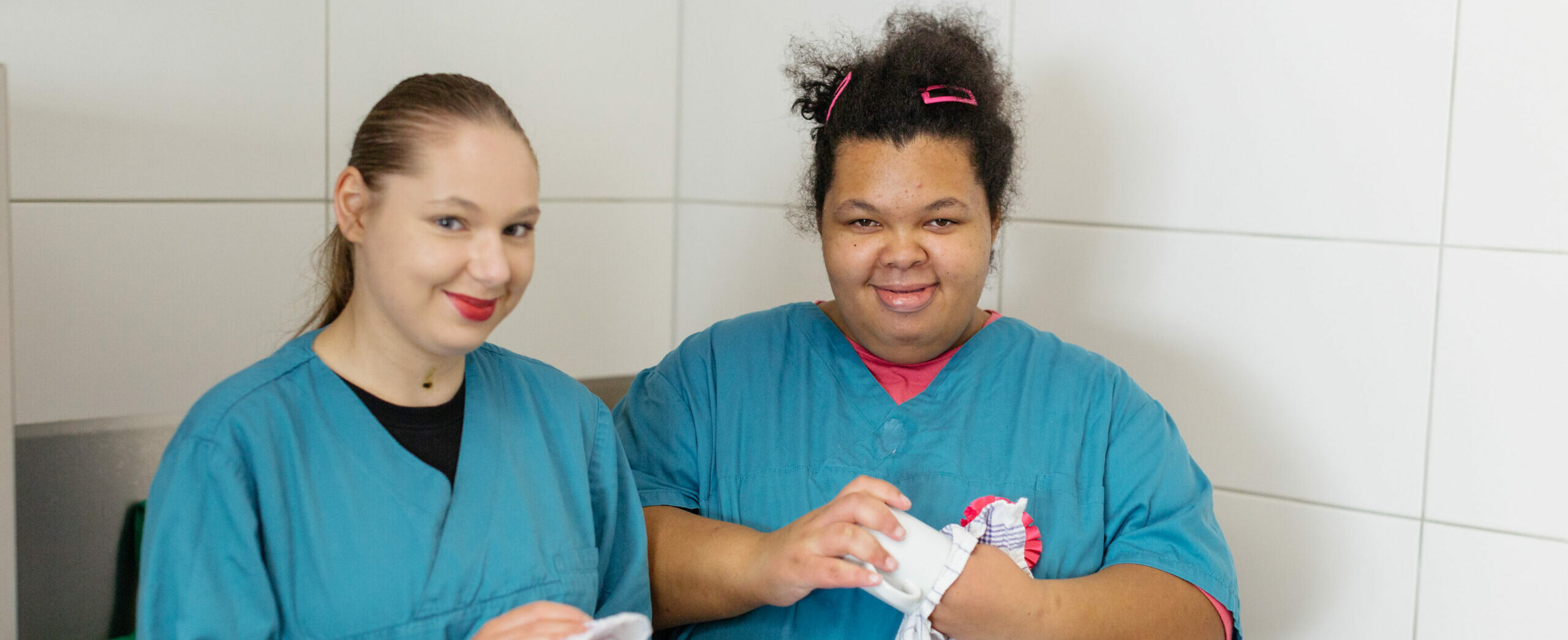 Zwei junge Frauen in einem Raum schauen freundlich in die Kamera. Eine junge Frau von ihnen trocknet eine Tasse ab. Sie arbeiten beim BBV Nord in der Hauswirtschaft.