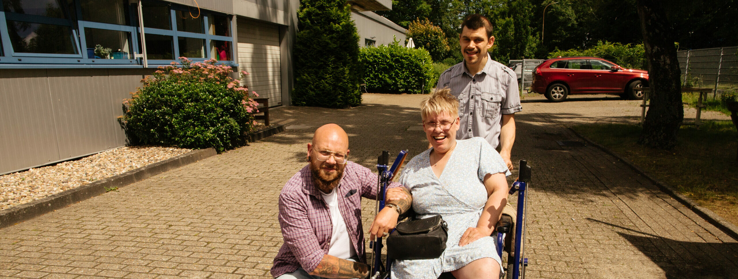 Draußen vor einer Werkstatt für Menschen mit Behinderungen sind drei Menschen auf dem Gelände vor der Kamera. Eine junge Frau sitzt in einem Rollstuhl. Ein Mann steht hinter der Frau. Ein Mann kniet sich links neben der Frau im Rollstuhl. Alle drei Menschen lächeln in die Kamera.