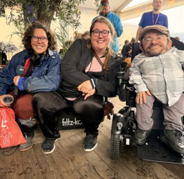 Anna-Helen Wippich vom Podcast „Kabelsalat“ sitzt gemeinsam mit Moderatorin Carola Schede (links) und dem Aktivisten Raúl Krauthausen (rechts) auf dem Watten Schlick Festival 2025 unter einem Pavillon. Alle drei lächeln in die Kamera. Im Hintergrund sieht man Menschen, Technik und das Zeltambiente des Festivals.